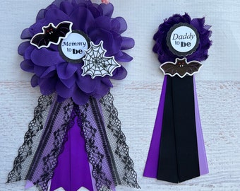May include: Two purple and black Halloween themed baby shower buttonholes. One says "Mommy to Be" with a bat and spider web. The other says "Daddy to Be" with a bat.