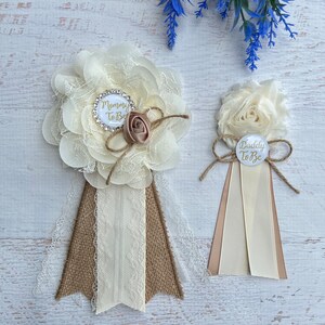 Może przedstawiać: Dwa białe i brązowe kwiatowe boutonnieres z wstążką z juty. Większy boutonniere ma białe k&oacute;łko z napisem "Mommy To Be" złotymi literami. Mniejszy boutonniere ma białe k&oacute;łko z napisem "Daddy To Be" złotymi literami.