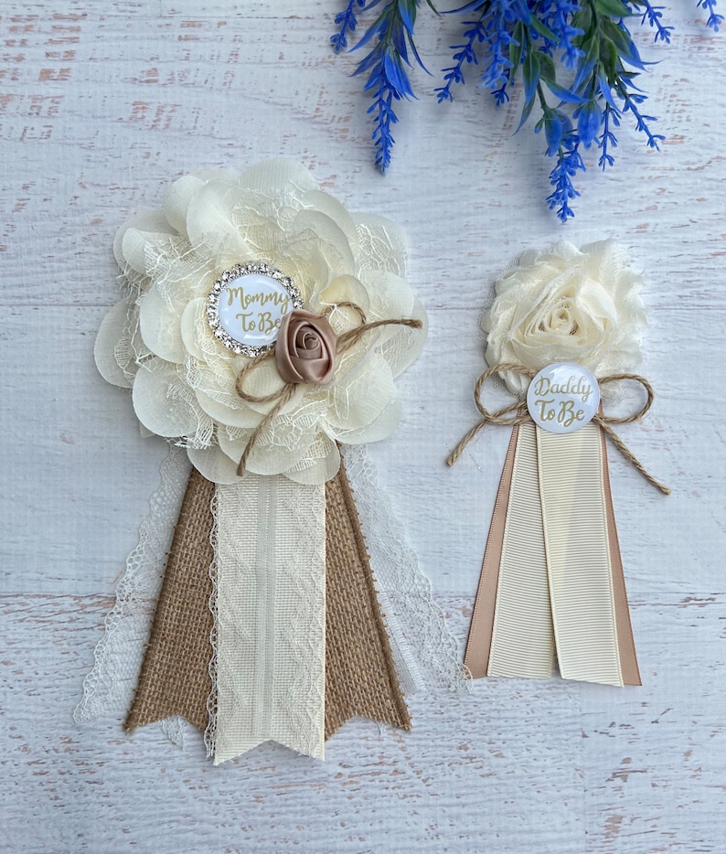 Może przedstawiać: Dwa białe i brązowe kwiatowe boutonnieres z wstążką z juty. Większy boutonniere ma białe k&oacute;łko z napisem "Mommy To Be" złotymi literami. Mniejszy boutonniere ma białe k&oacute;łko z napisem "Daddy To Be" złotymi literami.