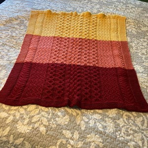 May include: A knitted throw blanket with a three-toned color gradient. The blanket is yellow, pink, and red. The blanket is made with a textured stitch pattern.