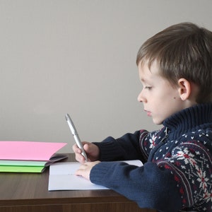 May include: A young boy wearing a blue sweater with a white snowflake pattern sits at a wooden desk, writing in a notebook with a silver pen. He has short brown hair and is looking down at his work. There are three notebooks stacked on the desk in front of him, one pink, one green, and one light green.