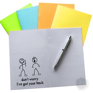 May include: A white notebook with a pen on top. The notebook has a drawing of two stick figures, one with a worried expression and the other with a confident expression. The text "don't worry I've got your back" is written below the figures.