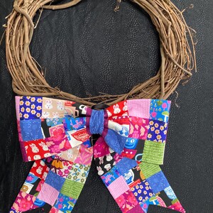 May include: A grapevine wreath with a large, colorful patchwork bow. The bow features a variety of fabrics with floral and bunny patterns in shades of pink, blue, red, and green. The wreath is a rustic, natural brown.