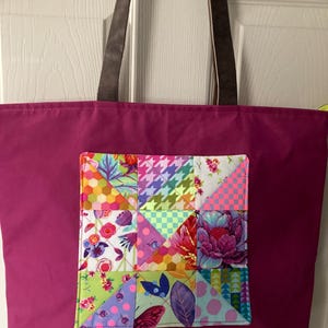 May include: A magenta tote bag with brown leather handles and a colorful patchwork pocket. The patchwork features floral, geometric, and houndstooth patterns in various colors. The bag is hanging on a white door.