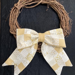 May include: A grapevine wreath with a large, decorative bow. The bow is made of fabric with a patchwork design in shades of gold and white. The wreath is a natural brown color.