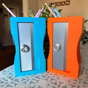 May include: Two pencil holders shaped like large manual pencil sharpeners, one in bright blue and one in vibrant orange. Each holder features a silver-colored rectangular front panel with a circular center detail and ribbed side edges.