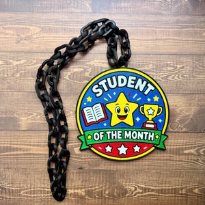 Puede incluir: Un medallón de premio circular Student of the Month con una estrella amarilla sonriente, un libro abierto y un trofeo dorado sobre un fondo azul, con el texto Student of the Month en letras blancas, unido a una gruesa cadena de plástico negro.