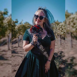 May include: A dark green velvet dress with a black sash that reads "Future Mrs. Gotnes". The bride is wearing heart-shaped sunglasses, a black veil, and holding a bouquet of red and black flowers. The background shows a vineyard.