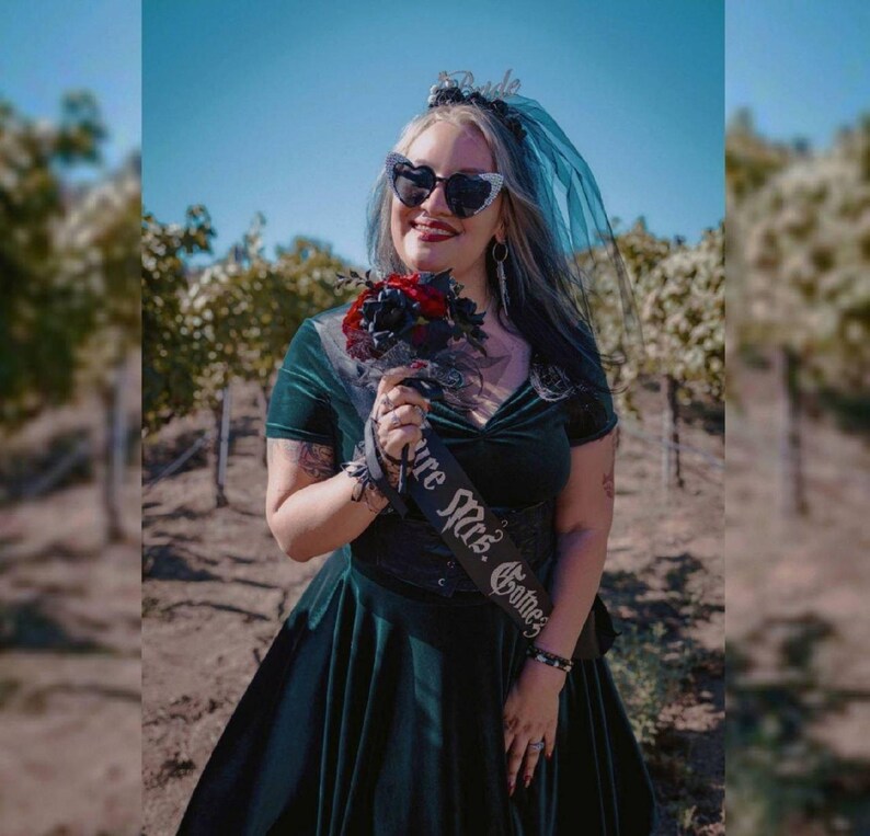 May include: A dark green velvet dress with a black sash that reads "Future Mrs. Gotnes". The bride is wearing heart-shaped sunglasses, a black veil, and holding a bouquet of red and black flowers. The background shows a vineyard.