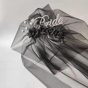 Op de afbeelding: Een zwarte bruidssluier met een decoratieve hoofdband. De hoofdband heeft het woord "Bride" in witte letters, zwarte rozen en imitatieparels. De sluier is gemaakt van transparante stof.