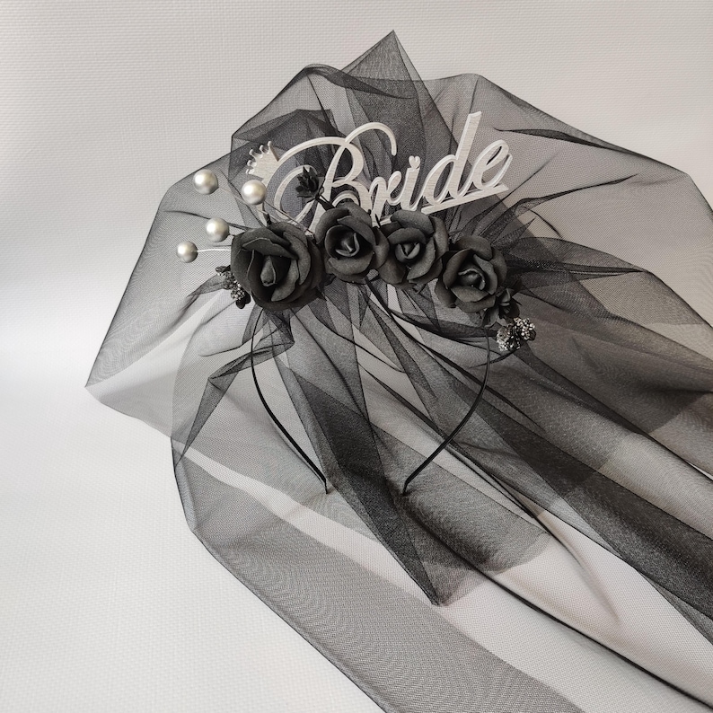Op de afbeelding: Een zwarte bruidssluier met een decoratieve hoofdband. De hoofdband heeft het woord "Bride" in witte letters, zwarte rozen en imitatieparels. De sluier is gemaakt van transparante stof.