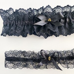 May include: Two black lace garters with satin bows and gold-colored stud accents. The top garter is wider and ruffled, while the bottom garter is a narrower band. Both are made of delicate, patterned lace.