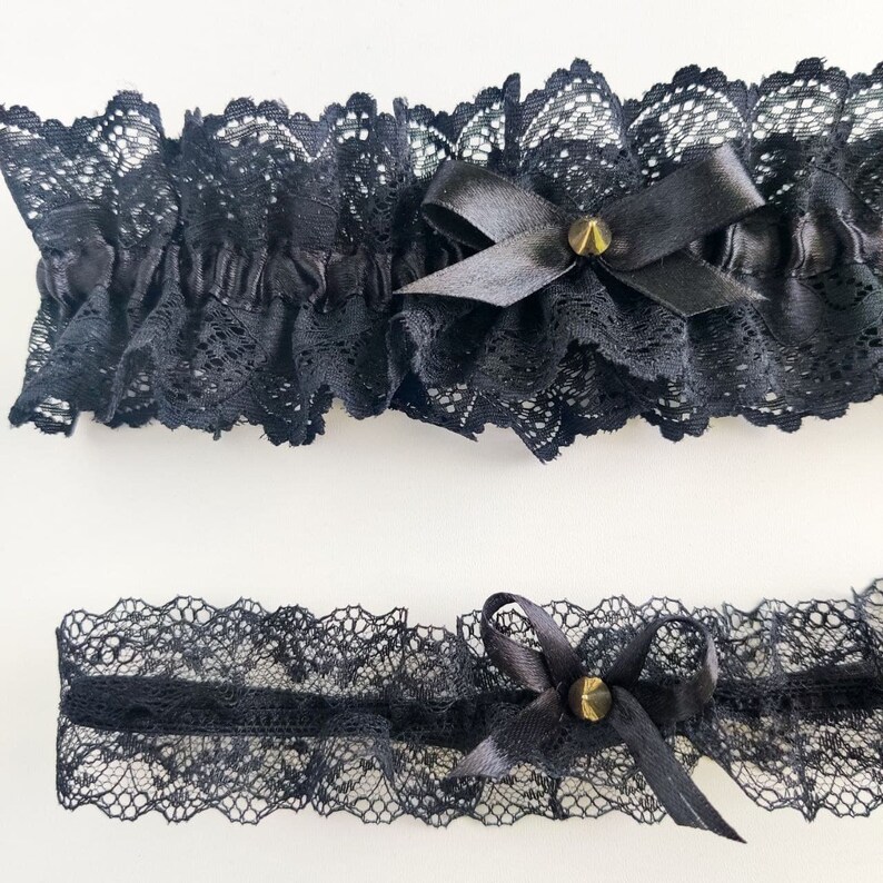 May include: Two black lace garters with satin bows and gold-colored stud accents. The top garter is wider and ruffled, while the bottom garter is a narrower band. Both are made of delicate, patterned lace.