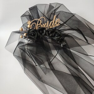 May include: Black bridal veil with a wooden "Bride" inscription and a crown detail. The veil features black fabric roses and gold-colored accents. The veil is made of sheer, flowing material.
