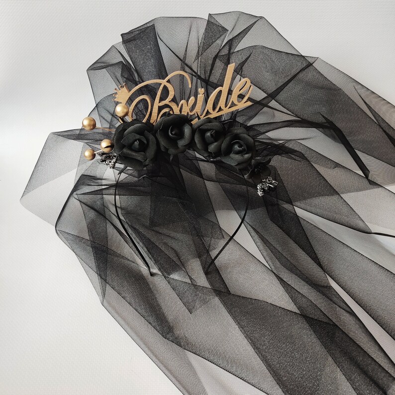 May include: Black bridal veil with a wooden "Bride" inscription and a crown detail. The veil features black fabric roses and gold-colored accents. The veil is made of sheer, flowing material.