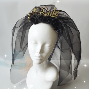 May include: A black bridal veil with a gold "Bride" headpiece. The headpiece features black roses, gold beads, and a gold crown. The veil is made of sheer black fabric.