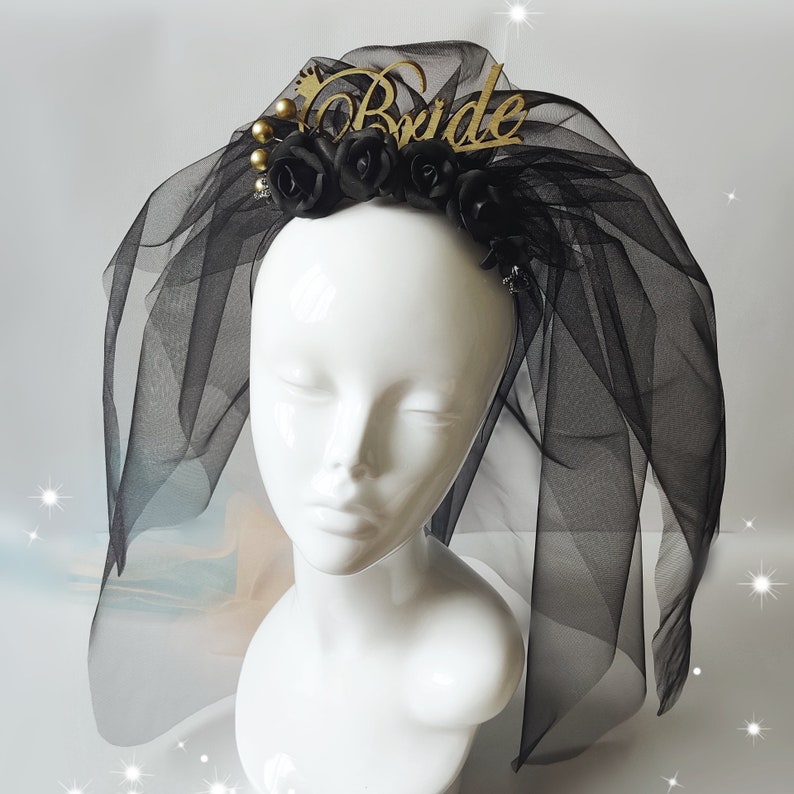 May include: A black bridal veil with a gold "Bride" headpiece. The headpiece features black roses, gold beads, and a gold crown. The veil is made of sheer black fabric.