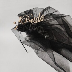 May include: Black bridal veil with a wooden "Bride" inscription. The veil is adorned with black roses and gold accents. The veil is made of a sheer, flowing fabric, perfect for a wedding or bachelorette party.