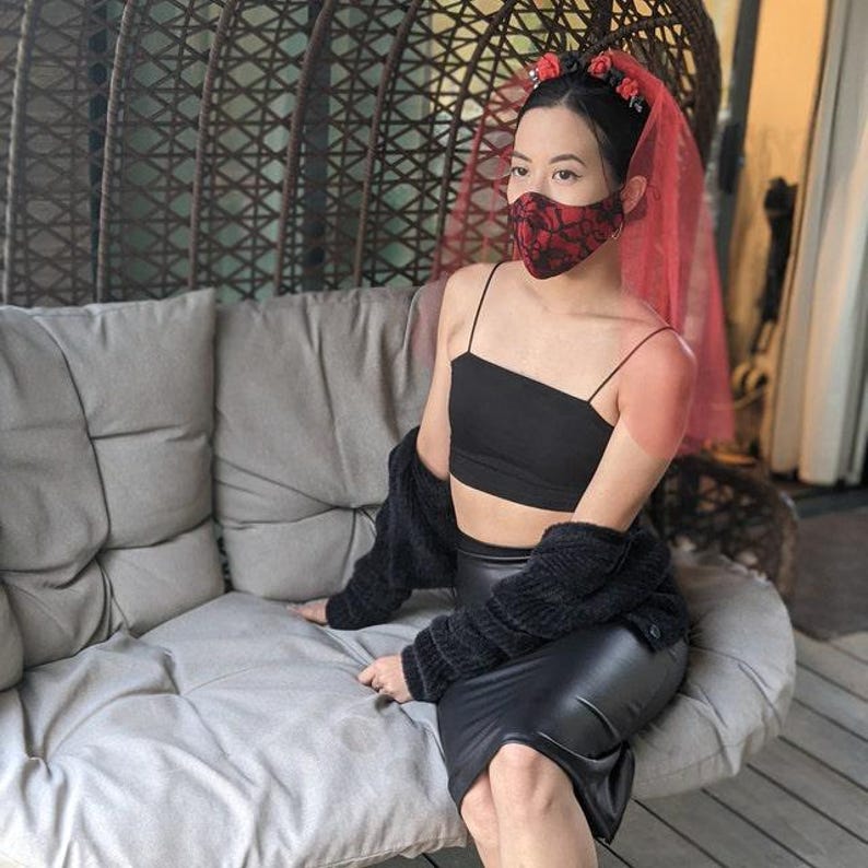 May include: A black crop top and a black leather skirt are paired with a black lace face mask and a red veil. A black cardigan is draped over the shoulders. The person is sitting on a gray cushion.