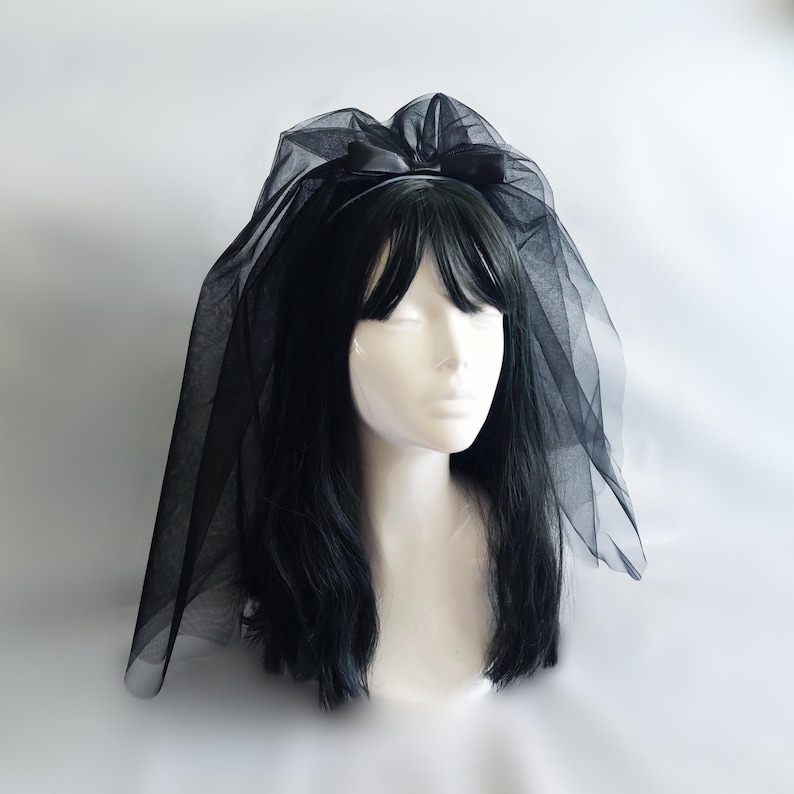 Op de afbeelding: Een zwarte tule sluier met een satijnen strik bevestigd aan een haarband. De sluier is gedrapeerd over een mannequinhoofd met zwart haar. Een bruids- of kostuumaccessoire.