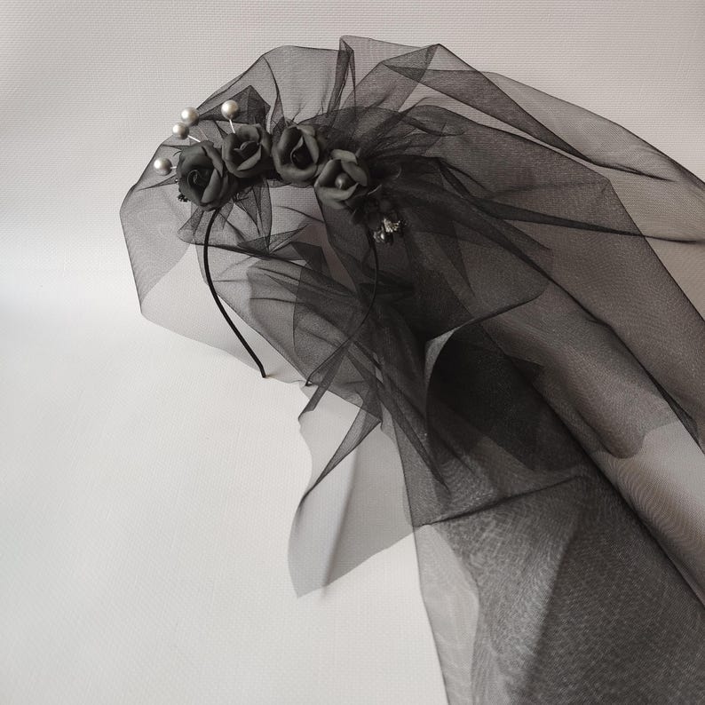 May include: A black tulle veil with a decorative headband. The headband features small black fabric roses and faux pearls. The veil is layered and draped, creating a soft, flowing effect. The veil is designed for special occasions.