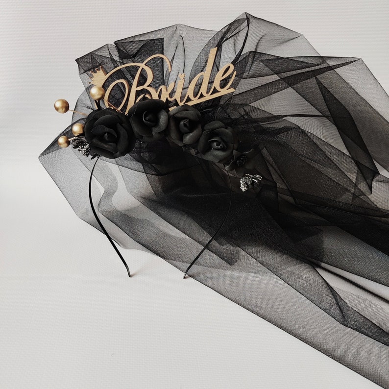 May include: A black tulle bridal veil with a headband featuring black roses, gold accents, and a wooden "Bride" inscription. The veil is layered and draped, creating a soft, flowing effect. The design includes a small gold crown.