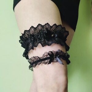 May include: Black lace garter set. The garters feature a scalloped lace design with satin bows and small gold-colored studs. The upper garter has a wider band and more elaborate detailing than the lower one. The background is a solid light green.