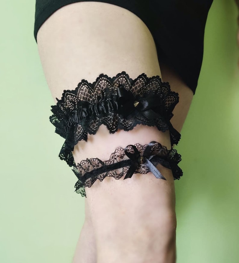 May include: Black lace garter set. The garters feature a scalloped lace design with satin bows and small gold-colored studs. The upper garter has a wider band and more elaborate detailing than the lower one. The background is a solid light green.