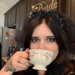 May include: A bride wearing a black veil and a floral crown with the word "Bride" in gold script. She is holding a white teacup with a floral pattern. The teacup has pink, yellow, and blue flowers.