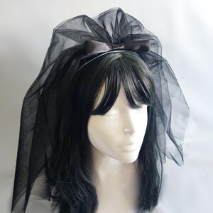Op de afbeelding: Een zwarte tule sluier bevestigd aan een haarband, met een satijnen strik. De sluier is gedrapeerd over een mannequinhoofd met zwart haar. Accessoire voor speciale gelegenheden.