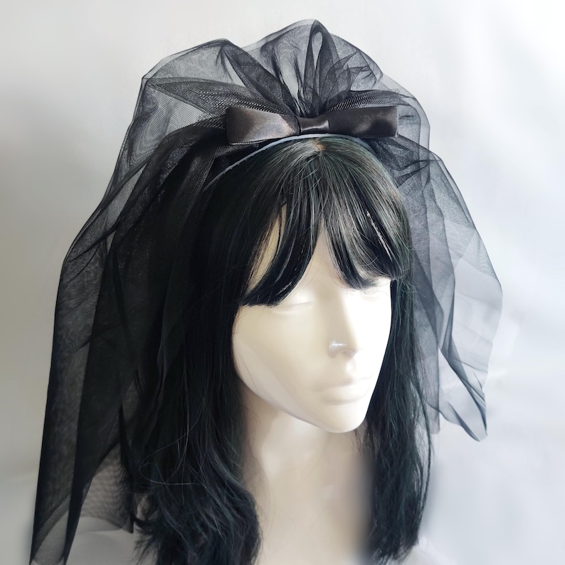 Op de afbeelding: Een zwarte tule sluier bevestigd aan een haarband, met een satijnen strik. De sluier is gedrapeerd over een mannequinhoofd met zwart haar. Accessoire voor speciale gelegenheden.