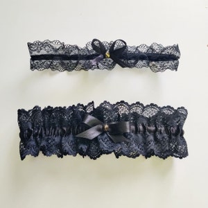 May include: Two black lace garters with satin bows and small gold-colored embellishments. The garters feature delicate scalloped edges and are designed for leg wear. The image is set against a plain white background.