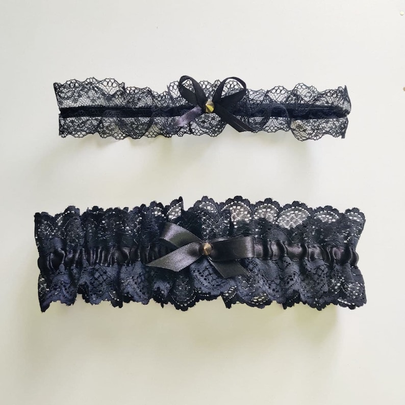 May include: Two black lace garters with satin bows and small gold-colored embellishments. The garters feature delicate scalloped edges and are designed for leg wear. The image is set against a plain white background.