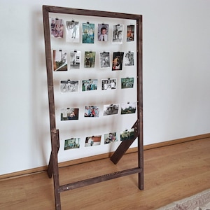 Può includere: Una cornice alta e rettangolare in legno marrone scuro appoggiata a terra, dotata di cinque fili orizzontali tesi all'interno. Numerose piccole fotografie rettangolari sono fissate a questi fili tramite piccole clip metalliche nere.