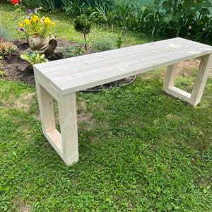 Könnte beinhalten: Eine helle Holzbank mit einer flachen, rechteckigen Sitzfläche und zwei stabilen, rechteckigen Beinen in Rahmenbauweise. Die Bank ist aus glatten, naturfarbenen Holzplanken gefertigt und zeigt ein klares, lineares Design mit sichtbarer Maserung.