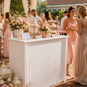 May include: A white rectangular wooden bar counter with a geometric panel design on the front. The top holds two gold tiered dessert stands, a large floral arrangement in a white vase, a smaller floral arrangement in a copper vase, and a sign.