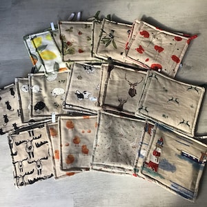 Könnte beinhalten: Eine Sammlung von 20 handgefertigten Topflappen mit verschiedenen Designs, darunter Tiere, Pflanzen und geometrische Muster. Die Topflappen sind aus natürlichem Leinenstoff gefertigt und haben einen beigefarbenen Hintergrund mit braunen Nähten.