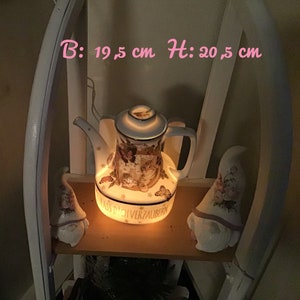 Könnte beinhalten: Eine weiße Porzellan-Teekanne mit floralem Design und dem Text "Lass dich verzaubern" auf der Unterseite. Die Teekanne ist von innen beleuchtet und hat einen warmen Schein. Die Teekanne ist 19,5 cm breit und 20,5 cm hoch.