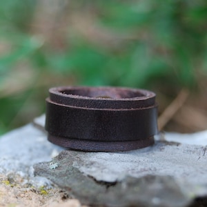 以下が含まれることがあります： シンプルなデザインの茶色の革のリング。リングは3層の革で作られており、岩の上に置かれています。