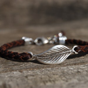 Könnte beinhalten: Ein braunes geflochtenes Lederarmband mit einem silbernen Blattanhänger.