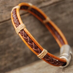 Puede incluir: Un brazalete de cuero trenzado en tonos marrones y tostados. El brazalete tiene un cierre magn&eacute;tico plateado.