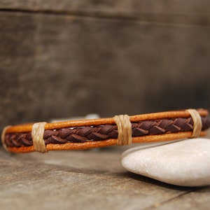 Puede incluir: Un brazalete de cuero marr&oacute;n con un dise&ntilde;o trenzado y detalles en cuero beige. El brazalete est&aacute; apoyado en una roca blanca.