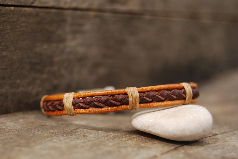 Puede incluir: Un brazalete de cuero marr&oacute;n con un dise&ntilde;o trenzado y detalles en cuero beige. El brazalete est&aacute; apoyado en una roca blanca.