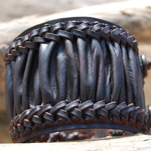 May include: A black leather bracelet with a braided design and a pleated texture. The bracelet is made of thick leather and has a rustic look.