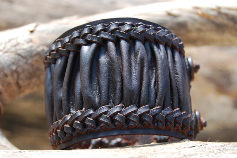 May include: A black leather bracelet with a braided design and a pleated texture. The bracelet is made of thick leather and has a rustic look.