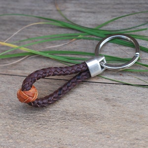 Op de afbeelding: Een bruine leren sleutelhanger met een gevlochten ontwerp en een kleine bruine leren bal. De sleutelhanger heeft een zilveren metalen ring.