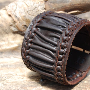 May include: A brown leather cuff bracelet with a braided design. The bracelet is made of thick, supple leather and has a rustic, handmade look.