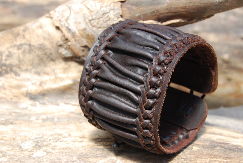 May include: A brown leather cuff bracelet with a braided design. The bracelet is made of thick, supple leather and has a rustic, handmade look.