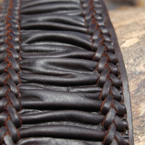 May include: Close-up of a brown leather belt with a braided design. The leather is textured and has a slightly wrinkled appearance.