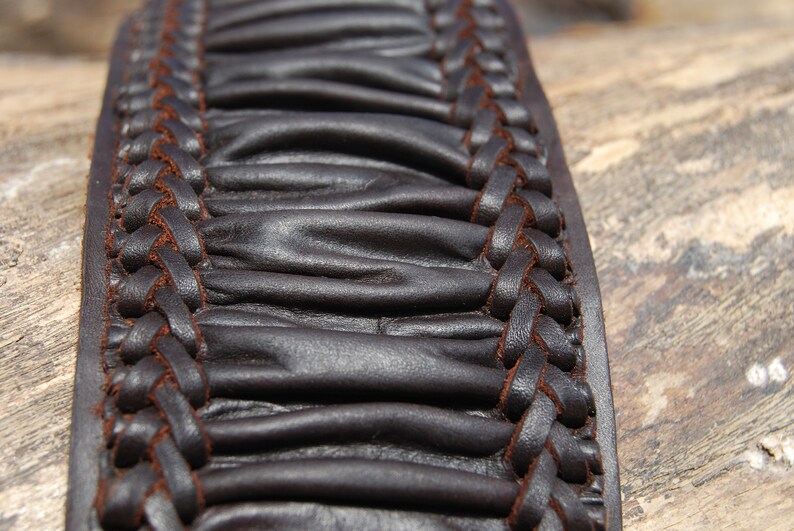 May include: Close-up of a brown leather belt with a braided design. The leather is textured and has a slightly wrinkled appearance.